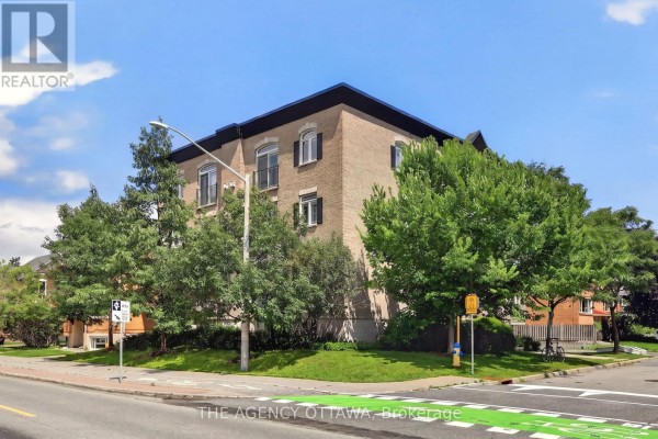 Corner view of the apartment building with trees, street signs, and a paved road in front. - 203 - 711 Churchill Avenue N, Ottawa