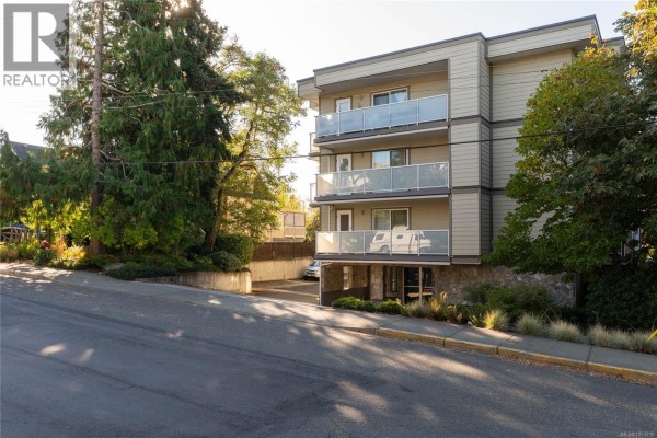 View of the apartment building from the street, showing entrance and parking. - 203 3230 Glasgow Ave, Saanich