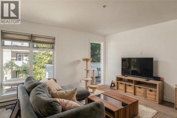 Living room featuring a sofa, television, and large window with tree views. - 203 3230 Glasgow Ave, Saanich