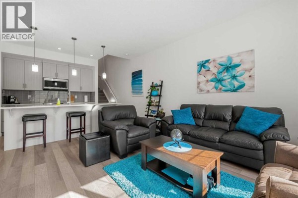 Open concept living area with grey kitchen island, black leather sofa, and wood plank flooring. - 203 Mahogany Grove Se, Calgary