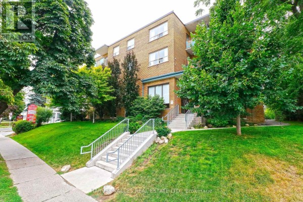 Side view of a brick apartment building with lush green trees and grass, and a paved sidewalk in front. - 204 - 1751 Victoria Pk Avenue, Toronto