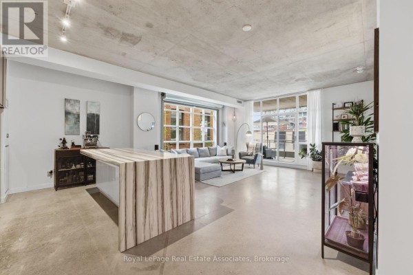 Open concept living area with concrete ceilings, island, sofa, and large windows. - 204 - 19 Brant Street, Toronto