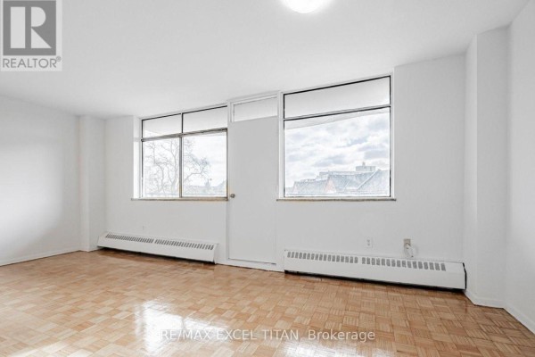 Living area with parquet flooring, two windows, and white walls. - 204 - 2255 Weston Road, Toronto