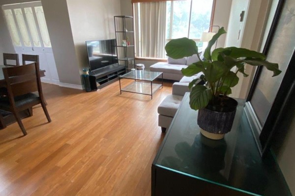 Living area with a sectional sofa, glass coffee table, and wood flooring, looking towards a large window. - 204 - 249 Major Mackenzie Drive E, Richmond Hill