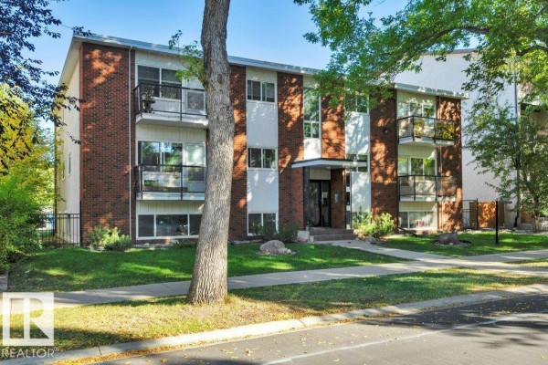 A wider exterior shot of the brick apartment building showing more of the surrounding lawn and trees. - #204 10540 80 Av Nw, Edmonton