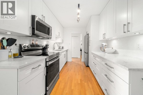White galley kitchen with white countertops, stainless steel appliances, and light wood floors. - 204 150 E 5th Street, North Vancouver