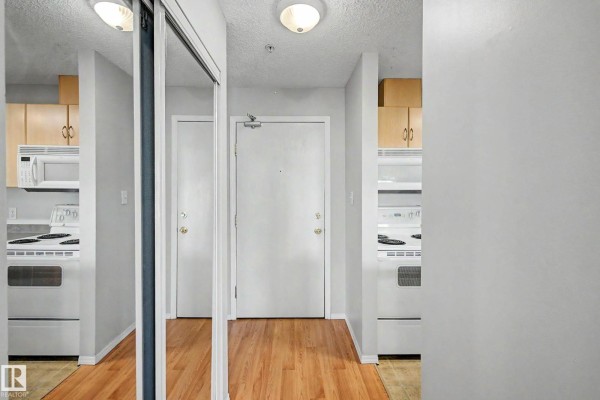 Hallway with mirrored closet doors, white doors, and entrance to kitchen with white appliances and wood floors. - #204 2430 Guardian Rd Nw, Edmonton