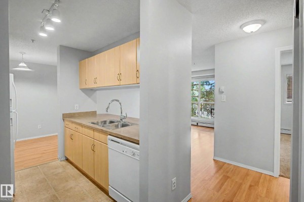 Kitchen with beige countertops, double sink, wood cabinets, white dishwasher, and view into the living room and hallway. - #204 2430 Guardian Rd Nw, Edmonton