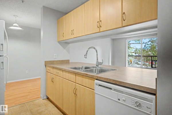 Kitchen with double sink, light wood cabinets, beige countertops, white dishwasher, and view of balcony with trees. - #204 2430 Guardian Rd Nw, Edmonton