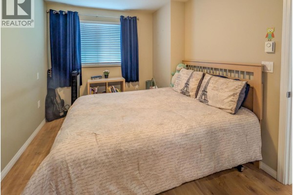 Bedroom with a large bed, wooden headboard, bedside table, and blue curtains covering the window. - 204 5000 Imperial Street, Burnaby