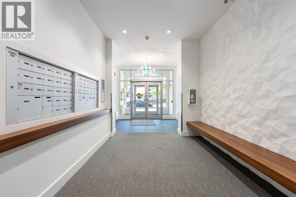 Apartment building lobby with mailboxes, textured wall, wooden bench, and glass doors. - 204 711 W 14th Street, North Vancouver