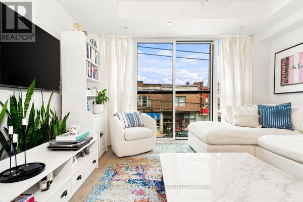 Living room with a large window, white sectional sofa, armchair, and bookshelf. - 205 - 899 Queen Street E, Toronto
