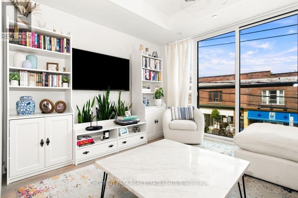 Living room with a large window, white sectional sofa, armchair, and entertainment unit. - 205 - 899 Queen Street E, Toronto