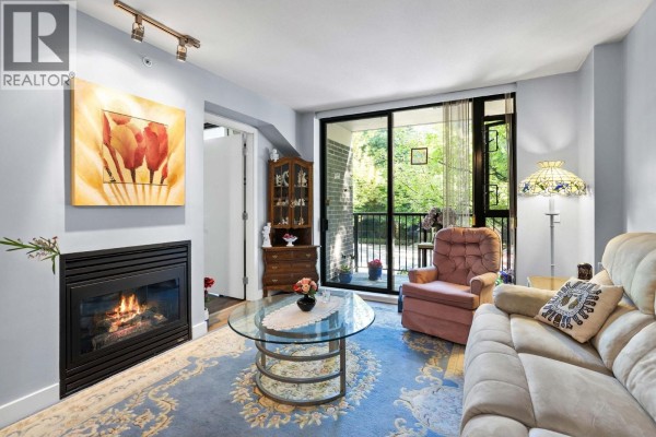 Living room with a gas fireplace, beige sofa, pink armchair, glass coffee table, and balcony access. - 205 175 W 1st Street, North Vancouver