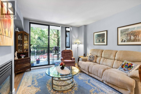 Living room with a fireplace, sofa, armchair, glass coffee table, and glass doors opening to a balcony. - 205 175 W 1st Street, North Vancouver