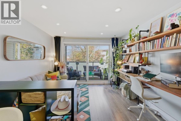 Living room with sofa, desk with computer, and wide wall shelves filled with books and plants. - 205 1864 Frances Street, Vancouver