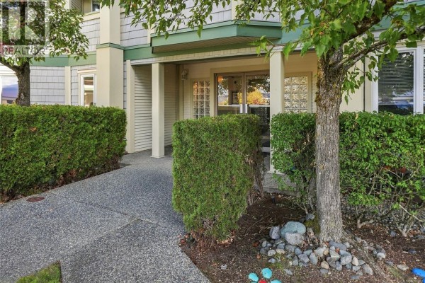 Covered entrance to apartment building with a sidewalk, hedges, and trees. - 205 4971 Songbird Pl, Nanaimo