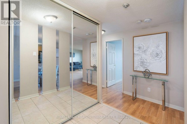 Hallway with mirrored closet doors, tiled floor, beige walls, a white door, and artwork. - 206 - 6720 Glen Erin Drive, Mississauga