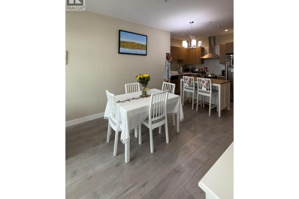 Dining area with a white table and chairs, next to a kitchen with wood cabinets and a breakfast bar. - 206 1838 Renfrew Street, Vancouver