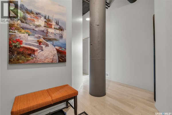 Hallway with light grey walls, an orange bench, a large painting, and a concrete column. - 206 211 D Avenue N, Saskatoon
