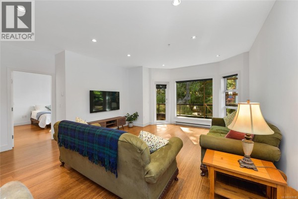 Living room with hardwood floors, two green sofas, a television, and a large bay window with a view. - 206 2589 Penrhyn St, Saanich