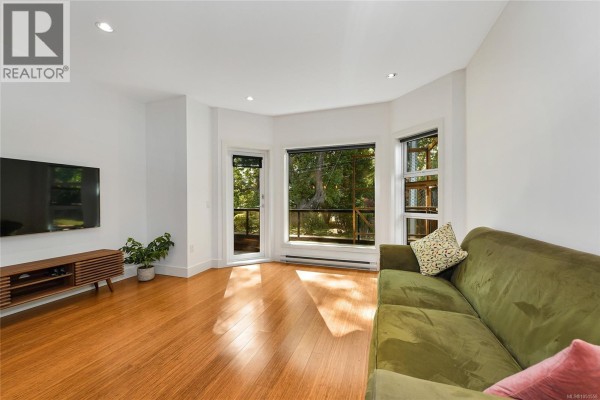 Living room with white walls, hardwood floors, a green sofa, and a large bay window overlooking trees. - 206 2589 Penrhyn St, Saanich