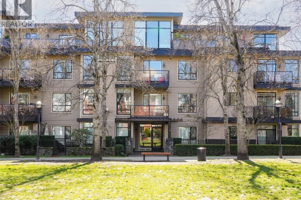 Living room with a sofa, fireplace, bookshelf, large windows, and beige carpet. - 206 330 Waterfront Cres, Victoria