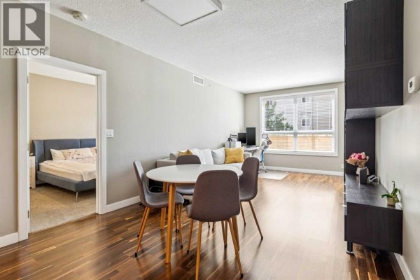 Living and dining area with dark wood floors, a dining table, and a glimpse into a bedroom. - 207, 1110 3 Avenue Nw, Calgary