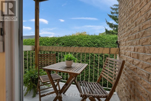 Small balcony with a wooden table, two chairs, and potted plants overlooking a hedge. - 207 - 190 Elgin Street W, Arnprior