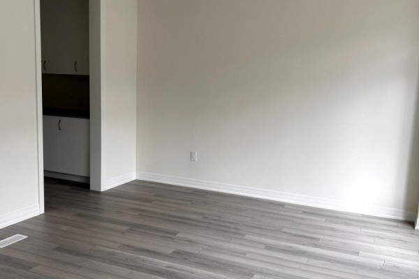 Empty room with light beige walls, light gray wood-look flooring, and a doorway to a small kitchenette. - 207 - 585 Colborne Street, Brantford