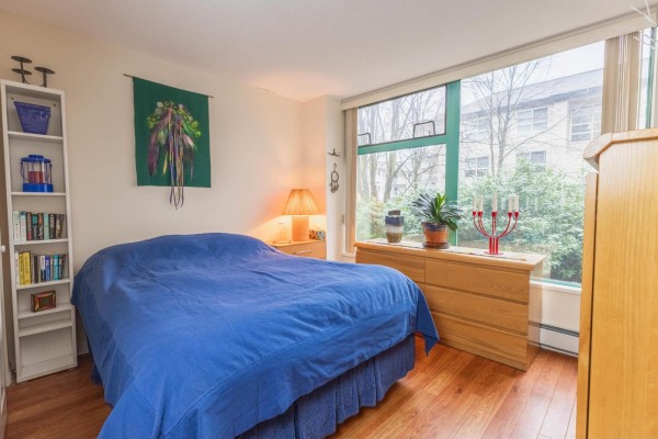 Bedroom with a blue bed, wooden dresser, and large window with trees outside. - 207 15466 North Bluff Road, White Rock