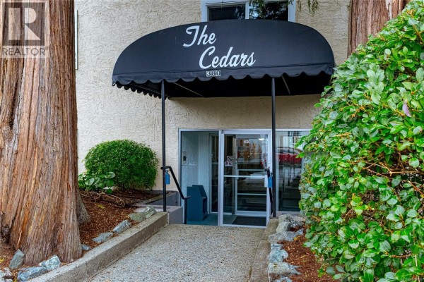 Entrance to "The Cedars" apartment building with a black awning and glass doors. - 207 380 Brae Rd, Duncan