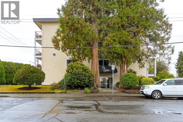 Front view of The Cedars apartment building with mature trees and a white SUV. - 207 380 Brae Rd, Duncan