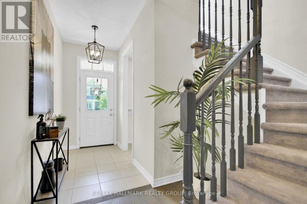 Interior hallway with beige walls, tile flooring, and a staircase with dark wood railing and carpeted steps. - 207 Willow Aster Circle, Ottawa