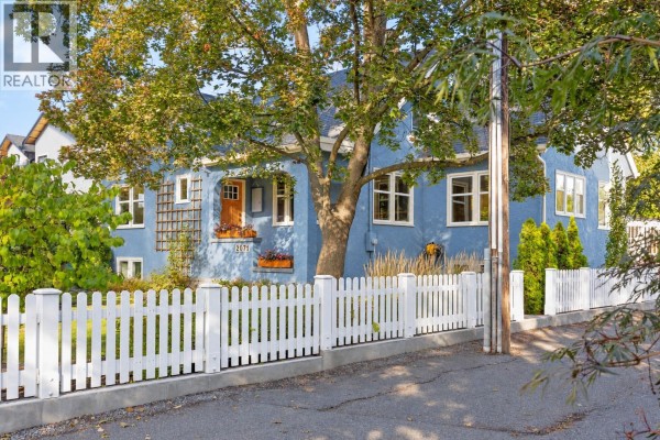 Front exterior of a light blue house with white picket fence and mature trees. - 2071 Abbott Street, Kelowna