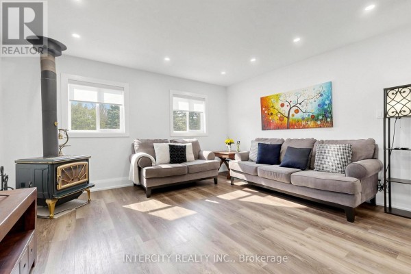 Living room with two grey sofas, a wood stove, two windows, and light wood-look flooring. - 2076 15th Side Road, New Tecumseth