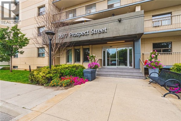 Close-up of the apartment building entrance with glass doors and landscaping. - 2077 Prospect Street Unit# 104, Burlington