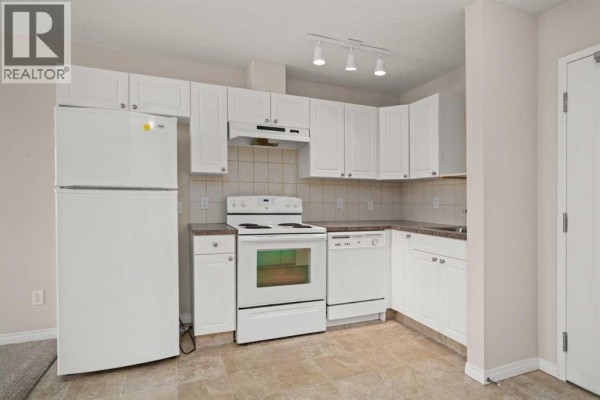 Apartment kitchen with white cabinets, white refrigerator, stove, dishwasher, and beige tile floor. - 208, 1010 Railway Street, Crossfield