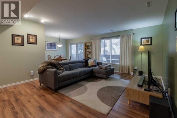 Living room with grey sectional sofa, coffee table, rug, and large windows with beige curtains. - 208, 2207 8 Avenue S, Lethbridge