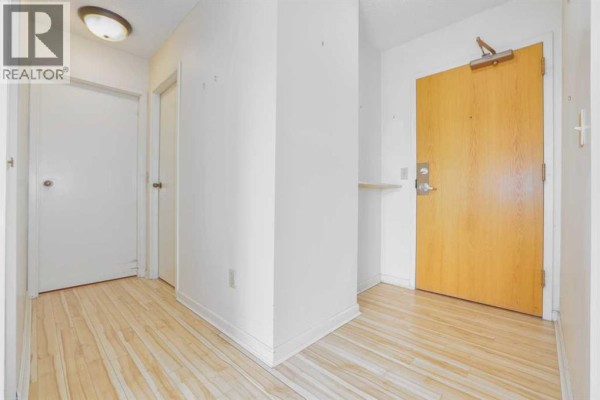Interior hallway with light wood floors, white walls, and a wooden front door. - 208, 80 Point Mckay Crescent Nw, Calgary