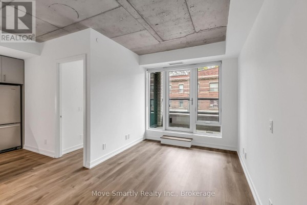 Living area with large window, wooden floors, and a view of brick buildings. - 208 - 115 Larchmount Avenue, Toronto
