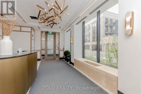 Apartment building lobby with a gold reception desk, modern chandelier, and large windows. - 208 - 123 Maurice Drive, Oakville