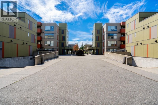 Driveway leading to apartment buildings with concrete retaining walls and green landscaping. - 208 12075 228 Street, Maple Ridge