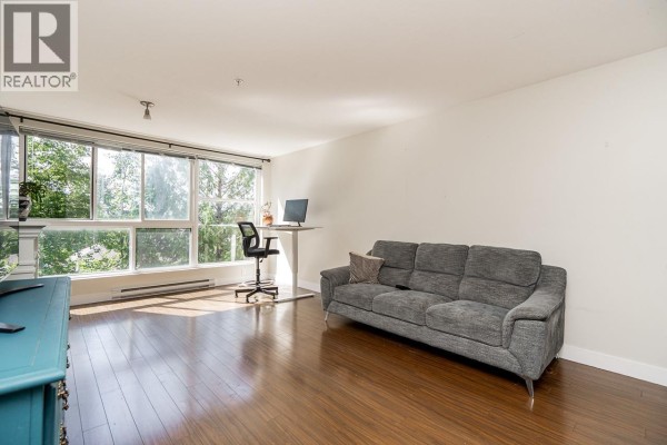 Living room with a gray sofa, desk with computer, and large window with tree views. - 208 12075 228 Street, Maple Ridge