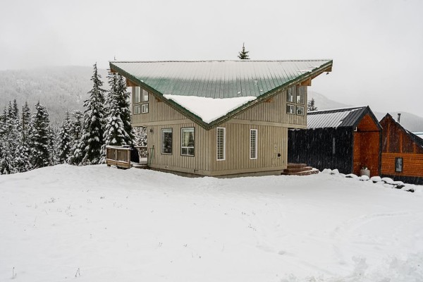 Living room with tan couch, armchair, wood stove, large window, and view of snowy trees. - 20806 Sakwi Creek Road, Agassiz