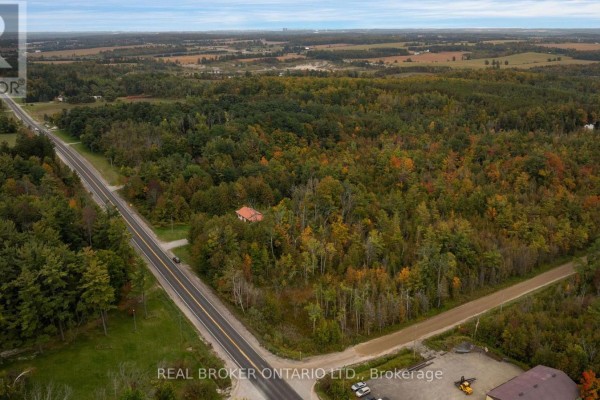 Aerial view of the property and surrounding landscape, featuring a road, wooded areas, and distant fields. - 2086 Penetanguishene Road, Springwater