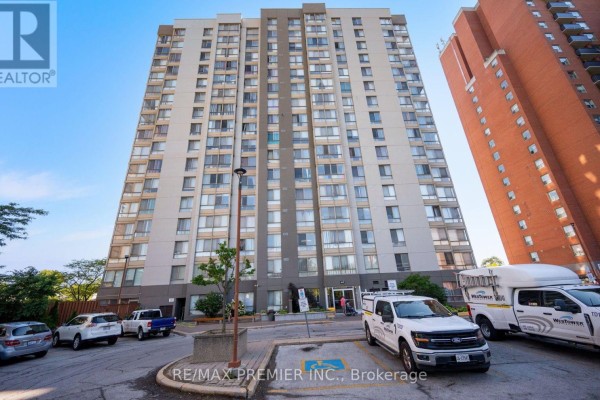 Low-angle view of an apartment building entrance with parked cars and trucks in the foreground. - 209 - 2470 Eglinton Avenue W, Toronto