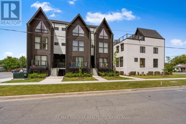 Exterior view of a modern townhouse complex with adjacent white building and landscaping. - 209 - 7277 Wilson Crescent, Niagara Falls