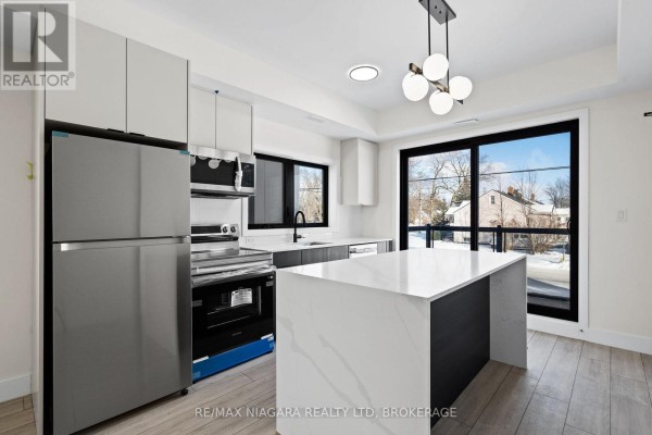 Bright modern kitchen with large window overlooking trees, white island, and stainless steel appliances. - 209 - 7277 Wilson Crescent, Niagara Falls