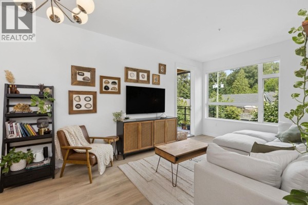 Living room with white sofa, armchair, media console, shelves, and balcony door visible. - 209 715 W 15th Street, North Vancouver